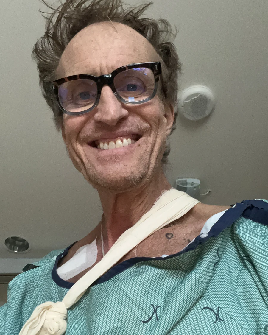 Jasen Rockliffe in a hospital gown and sling on a ward in Oakville, Ontario, grinning at the camera